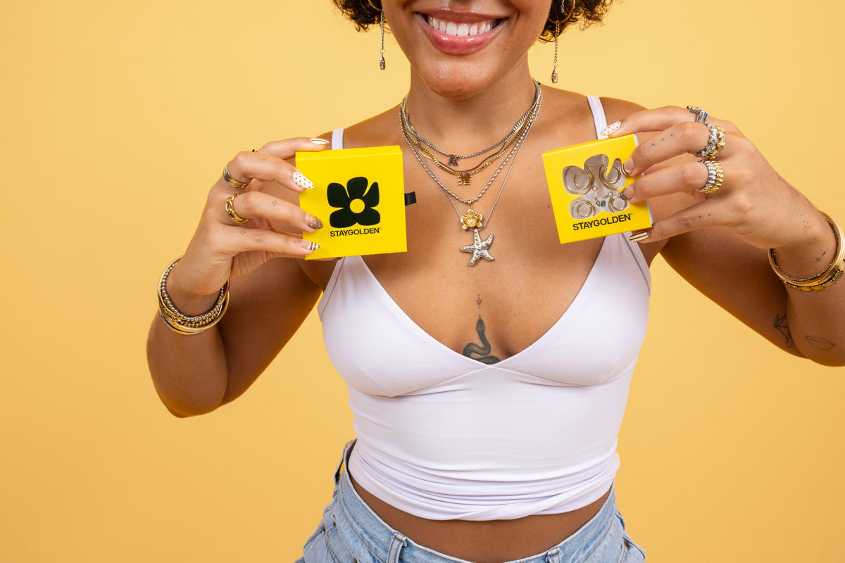 Layered Stay Golden necklaces, rings and bracelets on a model holding two yellow Stay Golden gift boxes